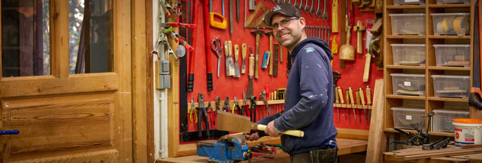 Jörg Klatt in der Tischlerwerkstatt mit einer Säge vor einer Werkzeugwand